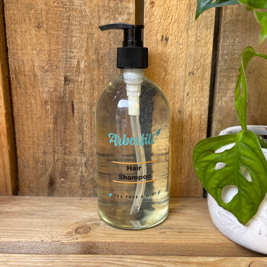 Clear shampoo bottle with pump on a wooden surface, surrounded by green plants.